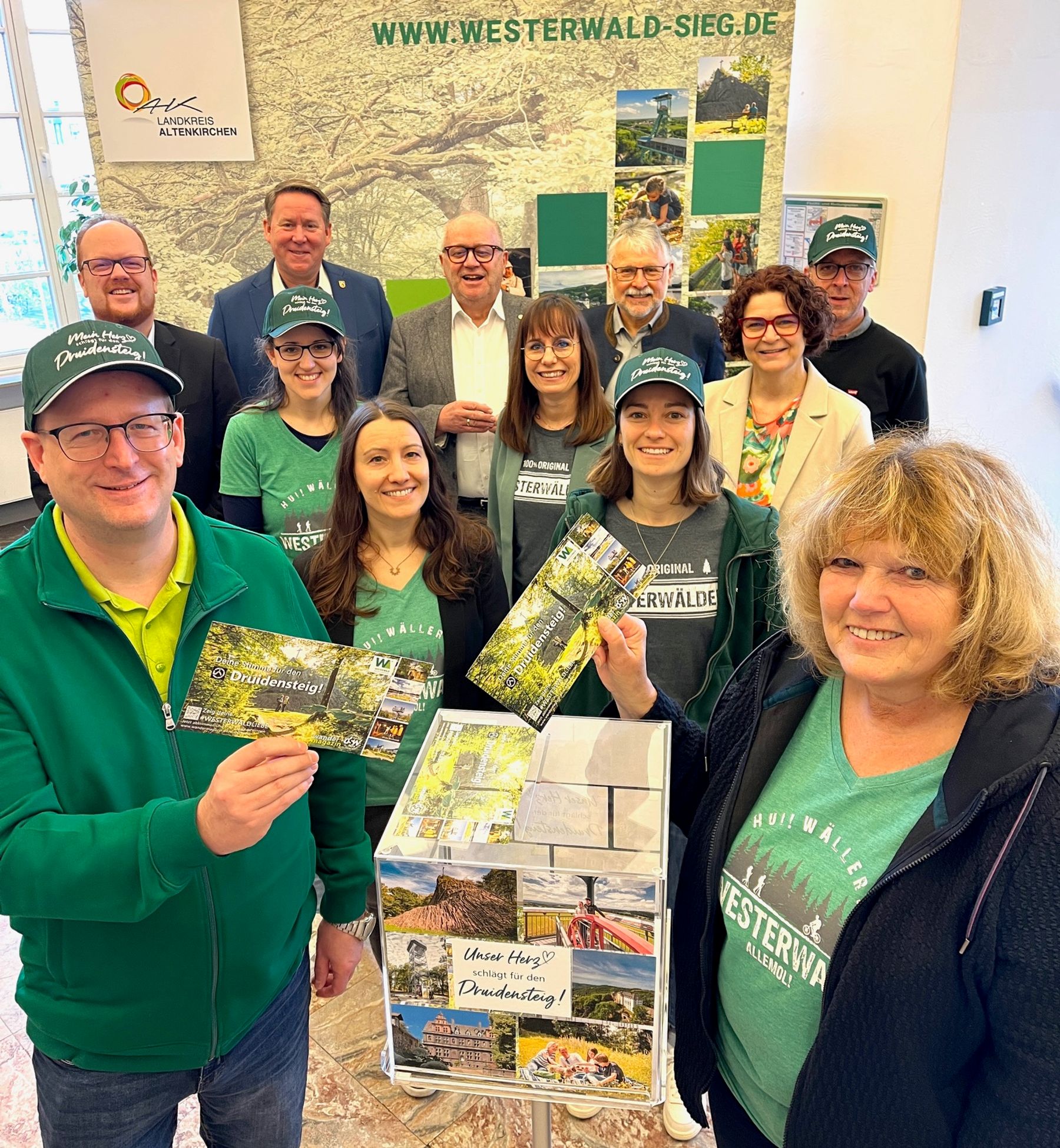 A group of people, some wearing green Westerwald shirts and hats, smile and hold brochures in front of a display box and a backdrop promoting Westerwald-Sieg tourism.